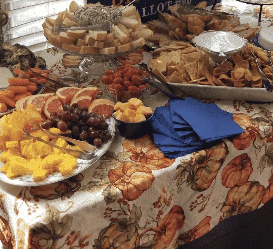 A table is set with an array of fruits, vegetables, crackers, and bread, all displayed on a fall-themed tablecloth.